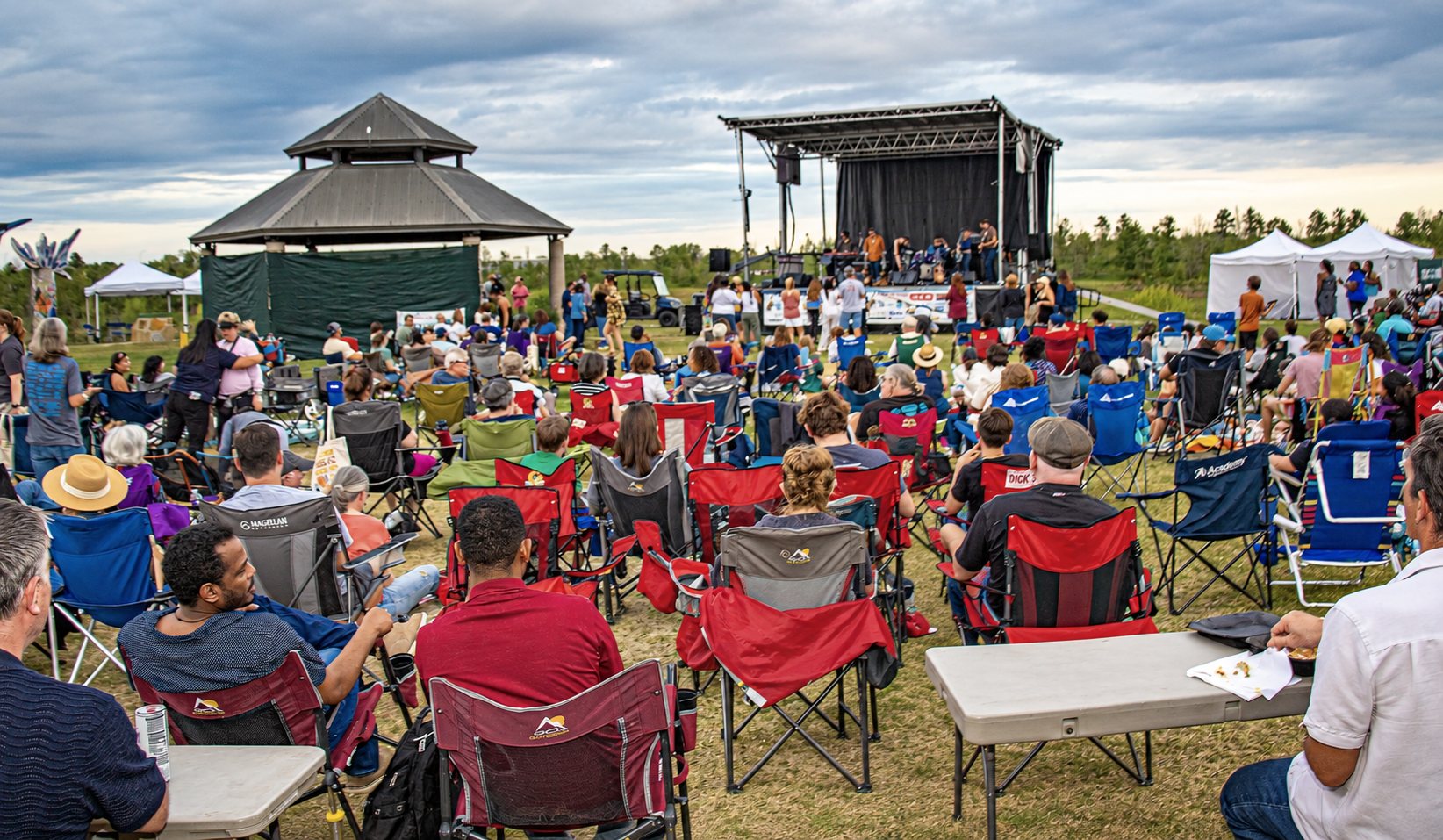 A crowd in lawn chairs facing a band on an outdoor stage at Willow Waterhole, with the gazebo and white tents nearby